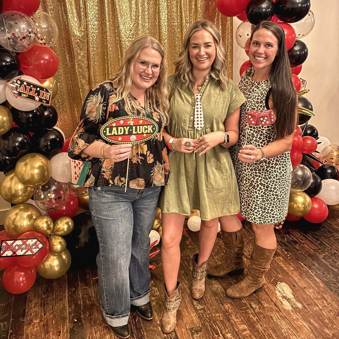 Three guests at the Casino Night photo booth holding a Lady Luck sign, dice, and a card-suit tie, cowboy boots and a gold backdrop